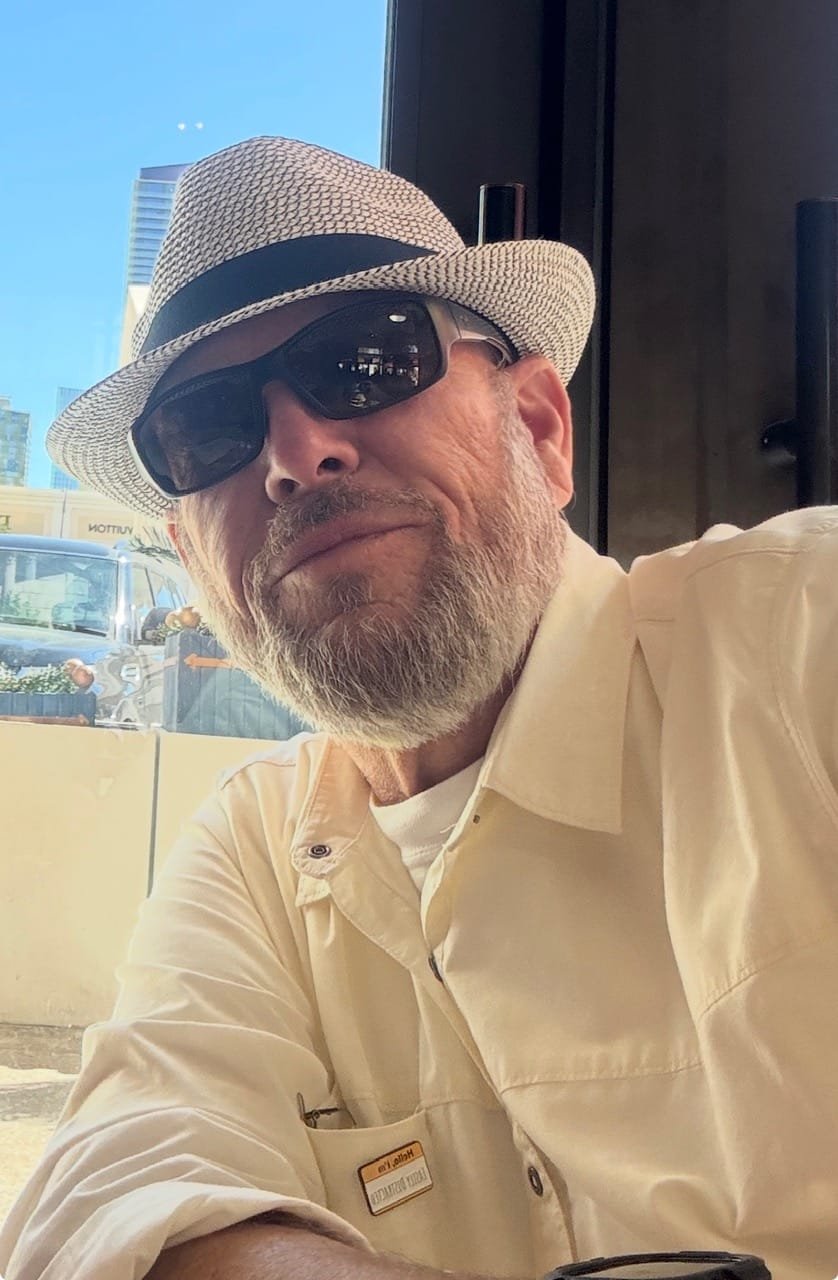 Hutch from JustOleHutch.com enjoying a sunny travel day, wearing a straw hat and sunglasses. He shares practical travel tips and budget advice for older travelers.